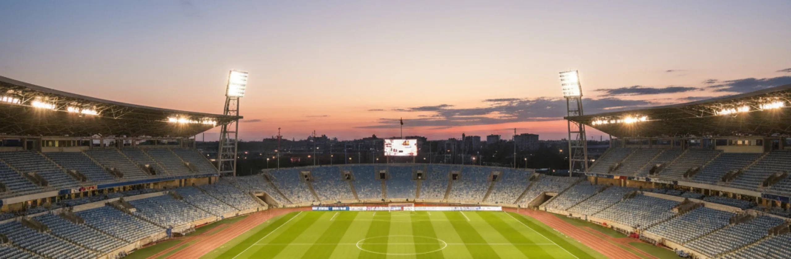 Empty stadium at dusk, floodlights lit above a green pitch ringed by a running track