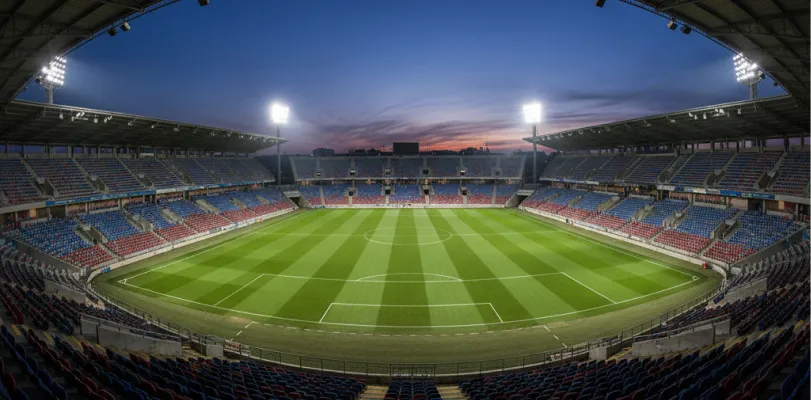 Empty floodlit stadium with red and blue seating around a striped grass pitch at dusk