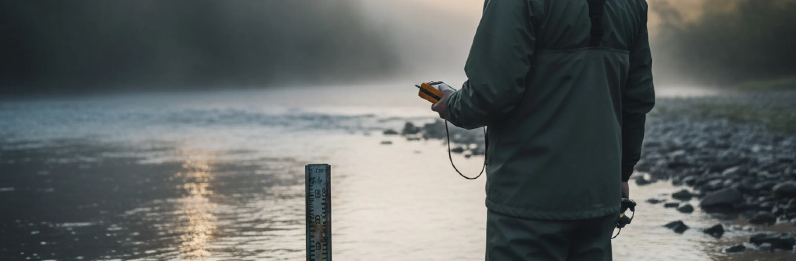 Researcher in a rain jacket holding a handheld meter beside a river gauge at dawn