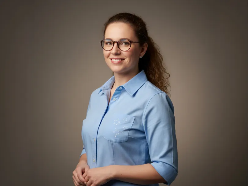 Dr. Yelena Marchetti, wearing glasses and a light blue button-down shirt, against a brown backdrop