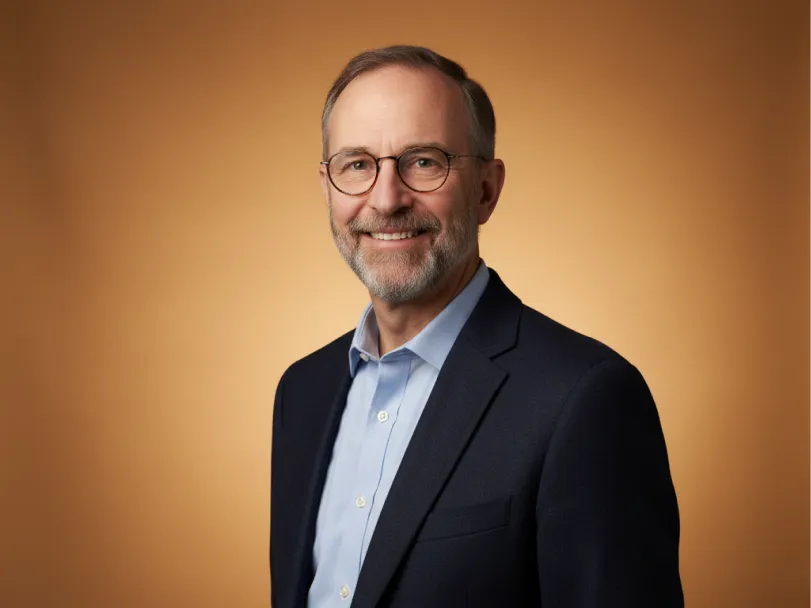 Elliot Braun in a dark jacket and light blue shirt, smiling against a warm amber backdrop