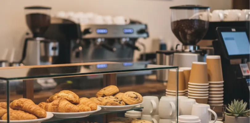 Pastries in a glass display case at a cafe counter, espresso machine and stacked cups behind