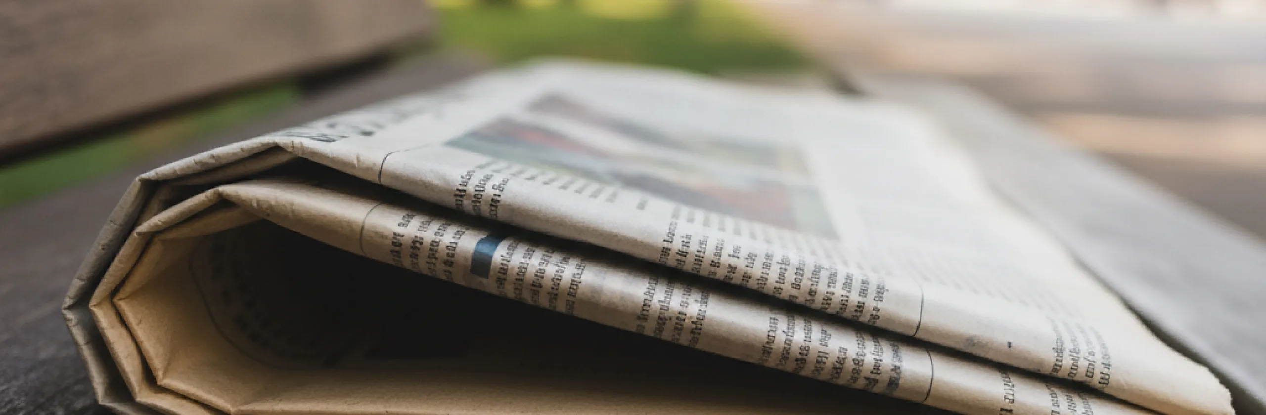 Folded newspaper lying on a wooden bench outdoors, columns of type and a color photograph