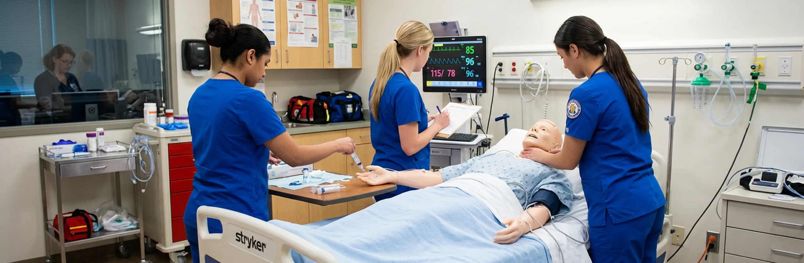 Three students in blue scrubs practice on a patient manikin beside a vital-signs monitor